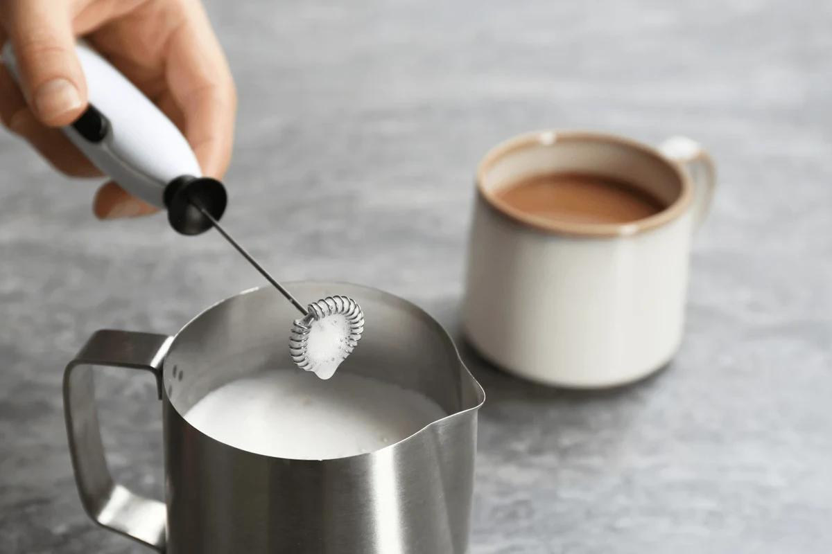 Electric milk frother being cleaned with warm water, soap, and a soft sponge in a kitchen sink