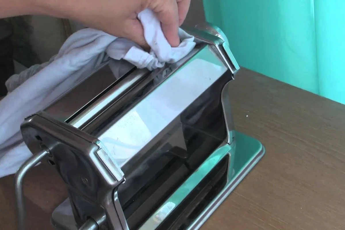 Hand cleaning a pasta machine with a brush and dry cloth on a kitchen counter