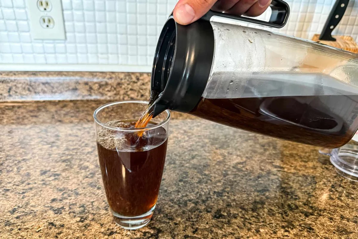 Glass of black cold brew coffee beside a kitchen counter and brewing jar