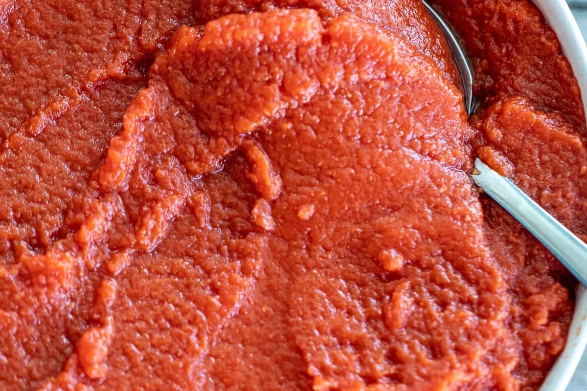 Bowl of tomato paste with spoon, garlic, herbs, and pasta ingredients on a kitchen counter