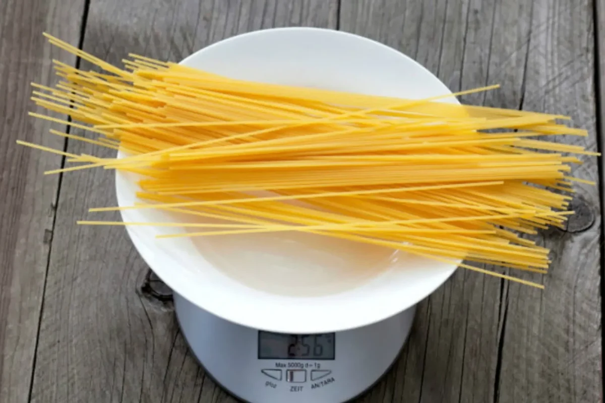Dry pasta in measuring cups beside a kitchen scale on a red kitchen counter