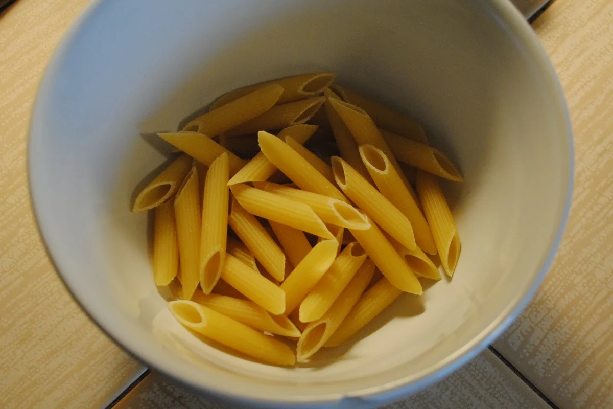 Measuring dry pasta in a cup beside a kitchen scale on a clean kitchen counter