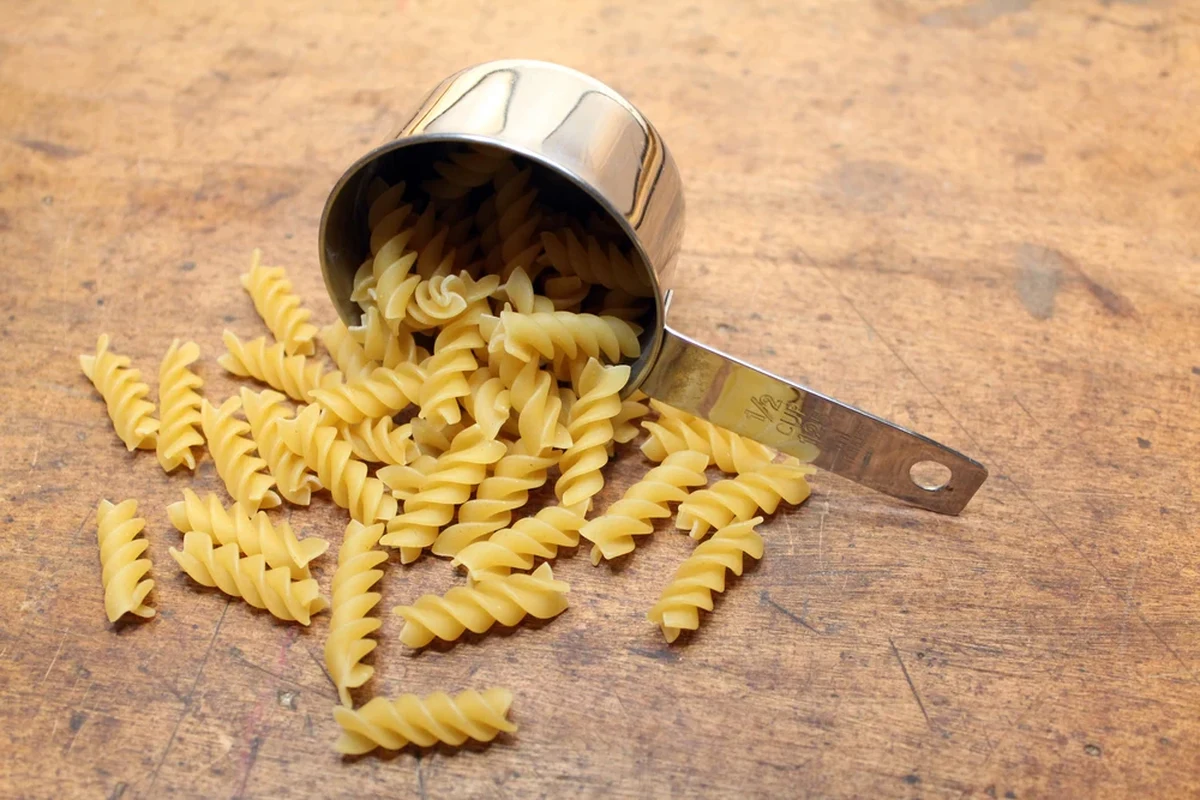 Dry pasta measured in a leveling cup beside a kitchen scale on a kitchen counter