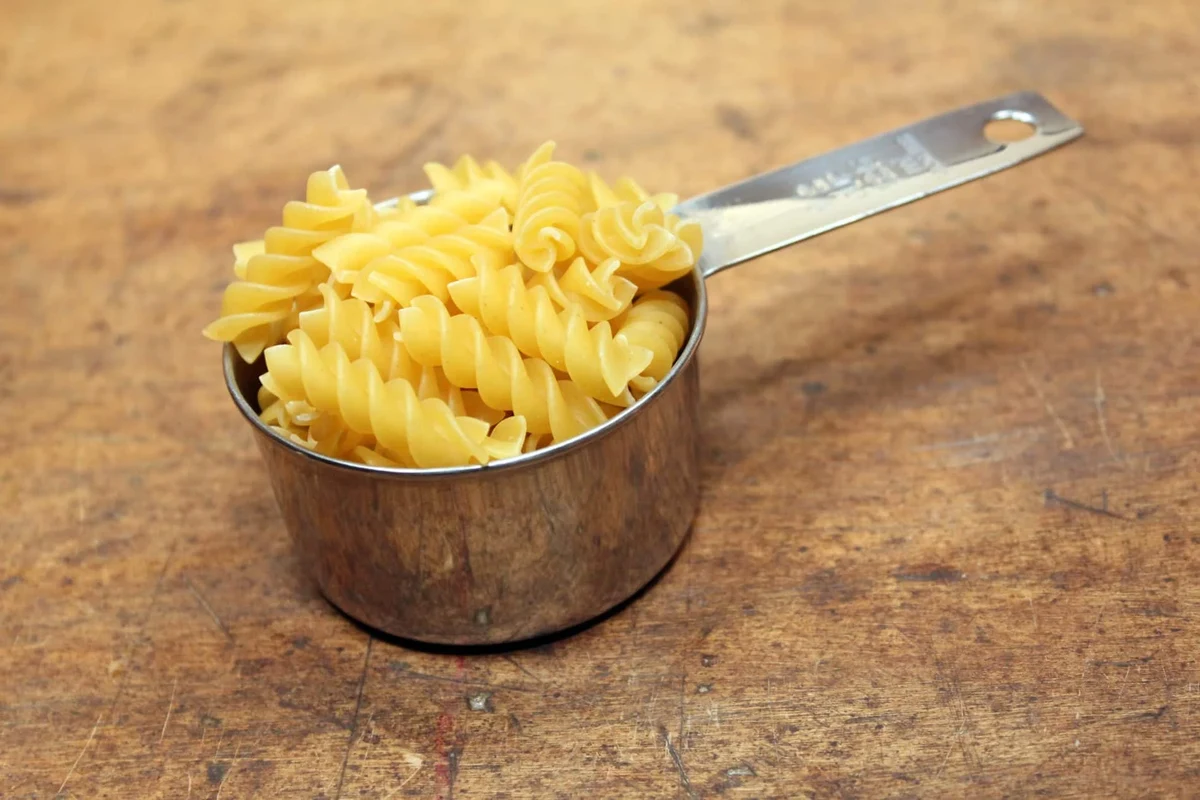 Measuring dry pasta in a cup beside a bowl of cooked pasta in a home kitchen