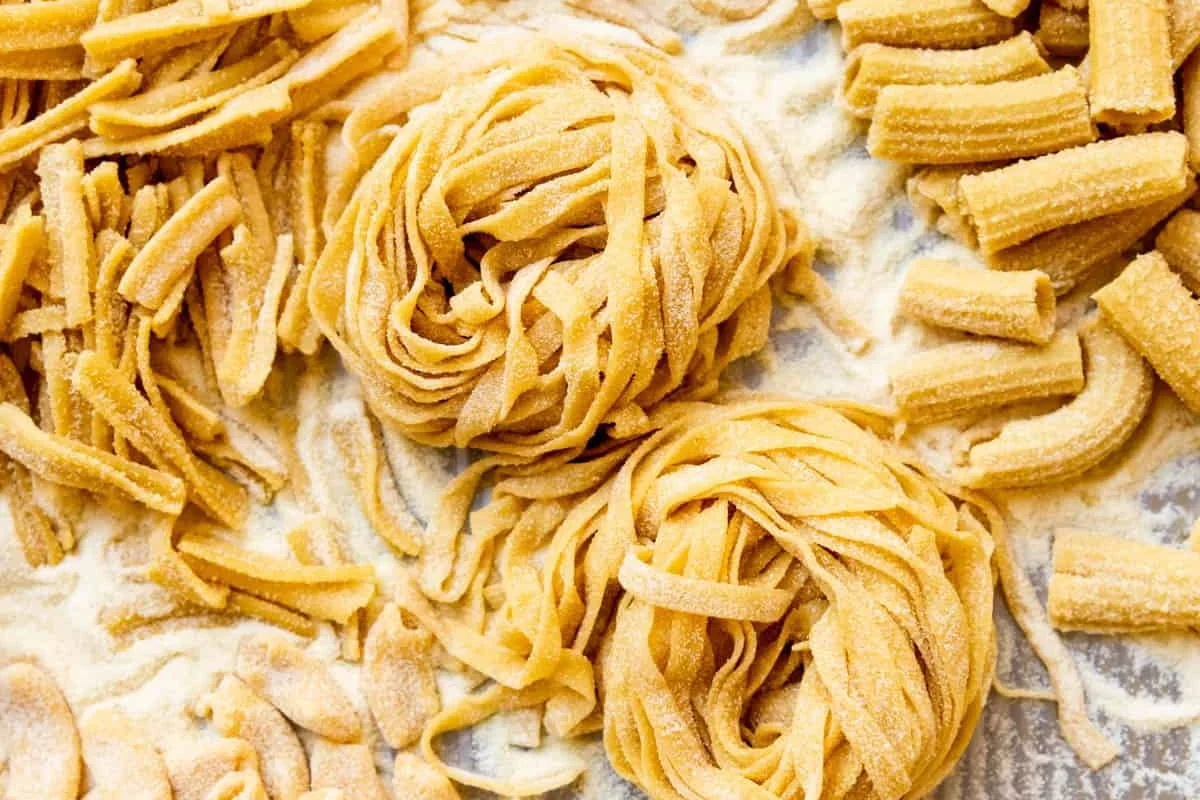 Ingredients for pasta dough with flour, eggs, water, and shaped noodles on a kitchen counter
