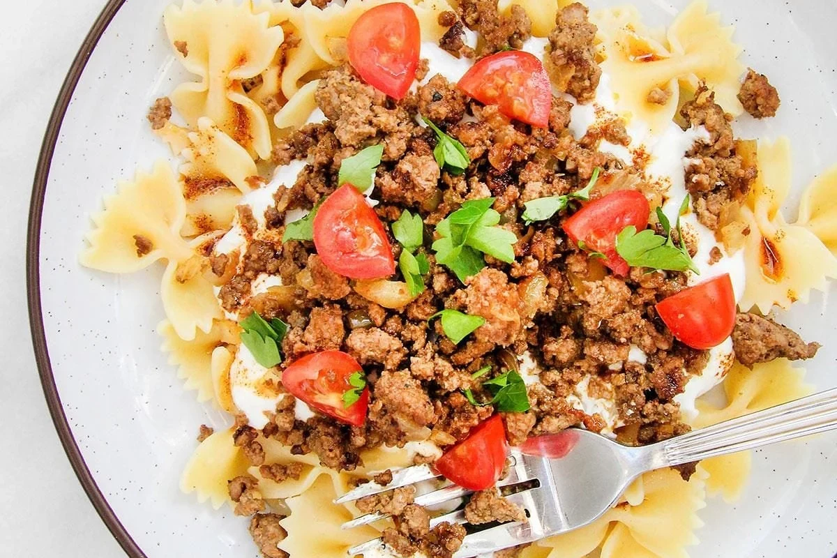 A bowl of Turkish-style pasta with tomato sauce, yogurt, herbs, and vegetables in a bright kitchen