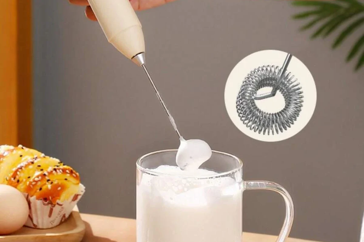 Electric milk frother foaming milk beside a home coffee setup in a kitchen